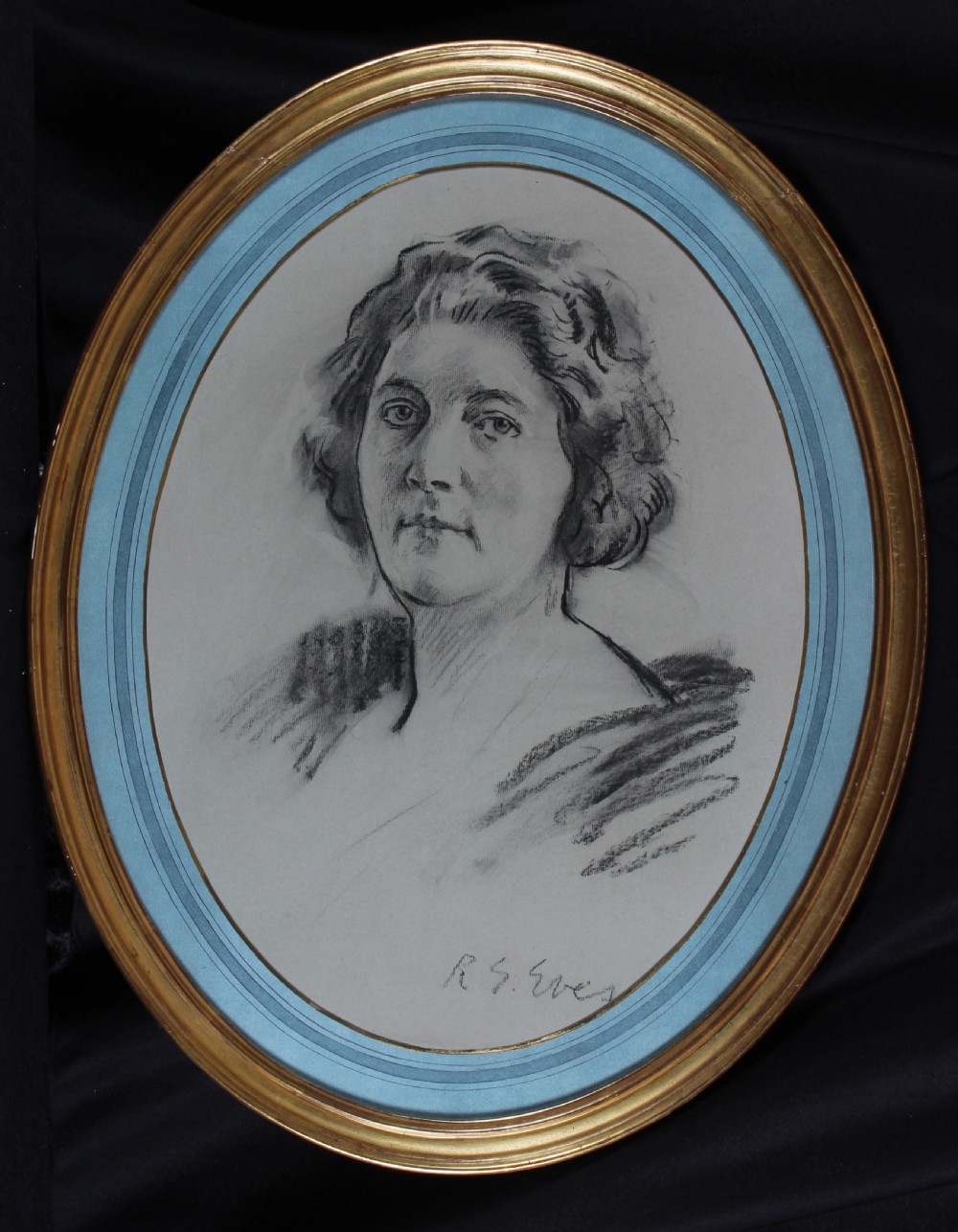 An oval gilt-framed charcoal portrait drawing of a woman signed by R. G. Eves set against a light blue mount.