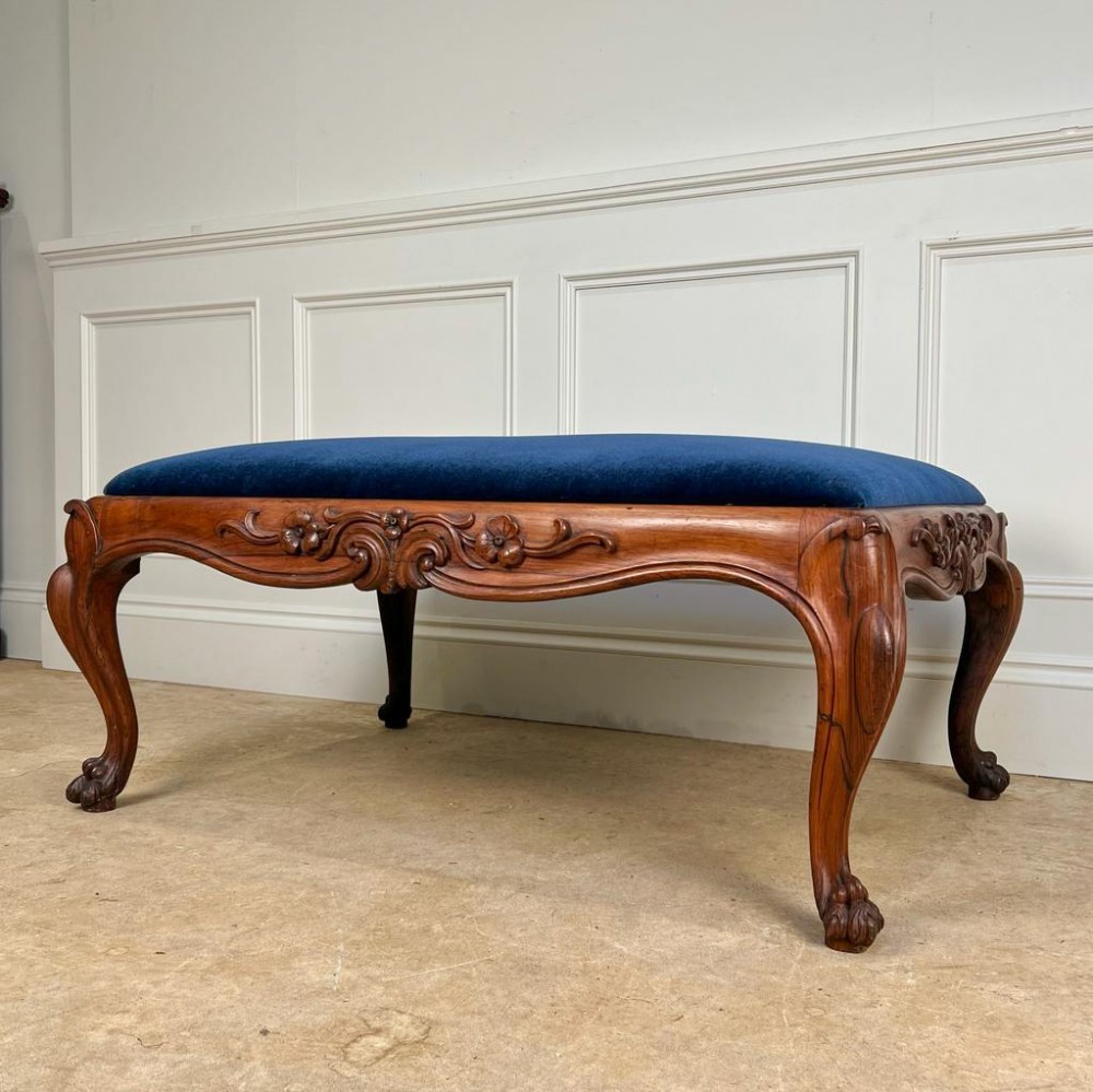 A large George IV rosewood ottoman stool with dark blue velvet upholstery, featuring carved floral aprons and cabriole legs.
