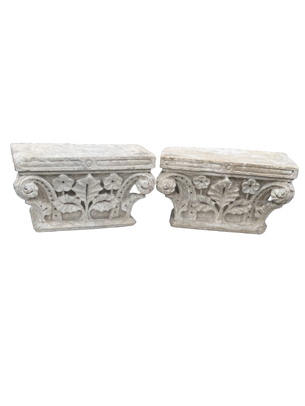 A pair of weathered, hand-carved grey stone capitals featuring floral and leaf motifs with scrolling volutes.