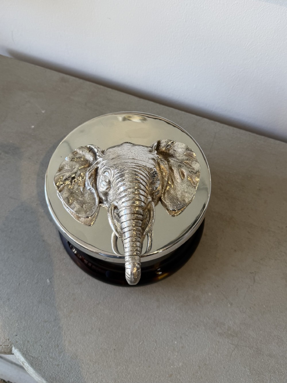 A circular faux tortoiseshell bowl with a polished silver-coloured metal lid featuring a sculpted elephant head in full relief.
