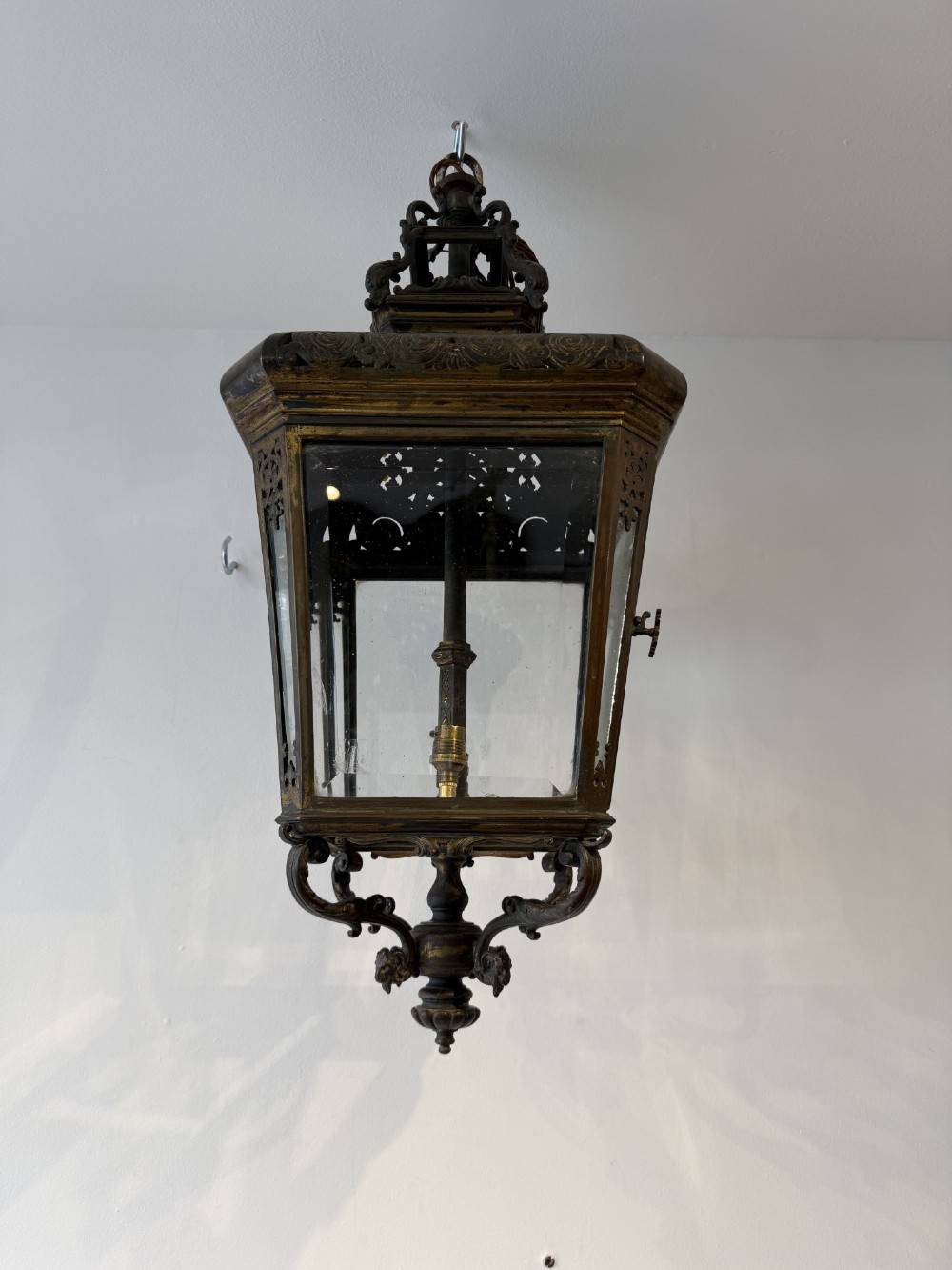 An antique patinated bronze hanging hall lantern featuring clear glass panels, ornate pierced metal cresting, and a scrolled lower finial.