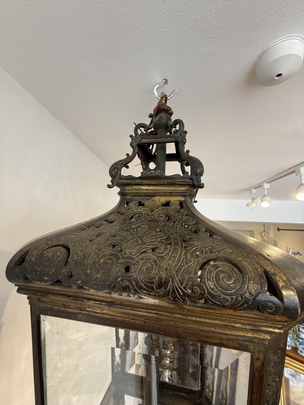 An antique patinated bronze hanging hall lantern featuring clear glass panels, ornate pierced metal cresting, and a scrolled lower finial. View III