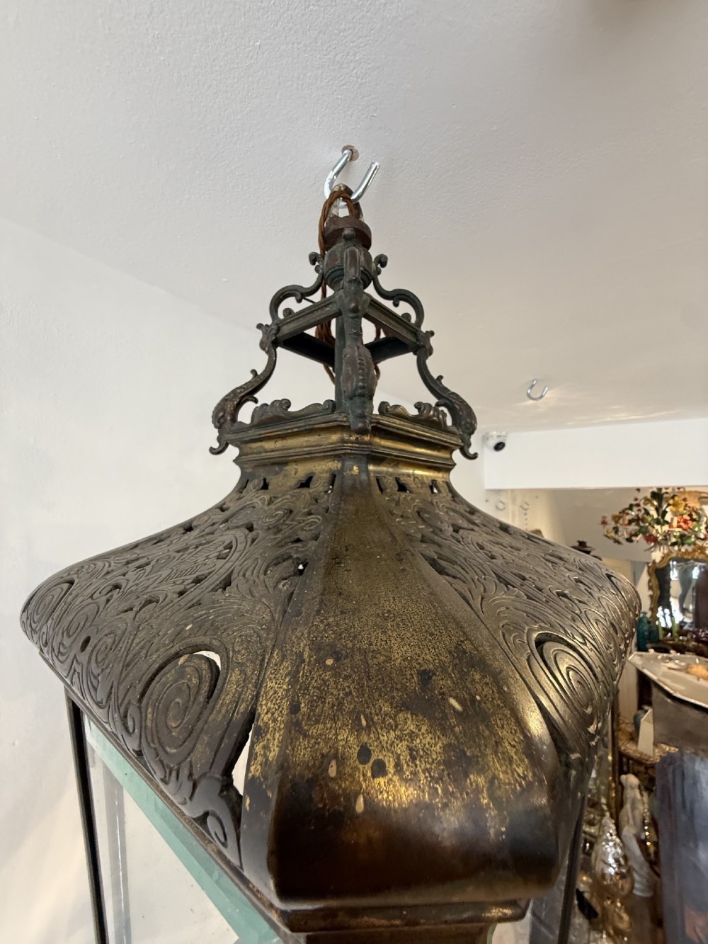 An antique patinated bronze hanging hall lantern featuring clear glass panels, ornate pierced metal cresting, and a scrolled lower finial. View IV