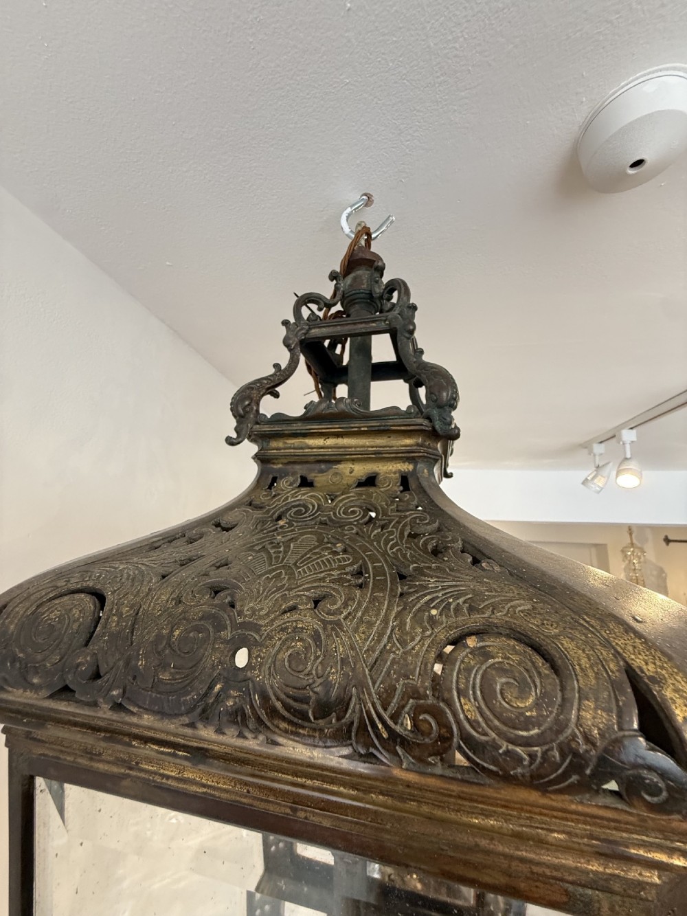 An antique patinated bronze hanging hall lantern featuring clear glass panels, ornate pierced metal cresting, and a scrolled lower finial. View V