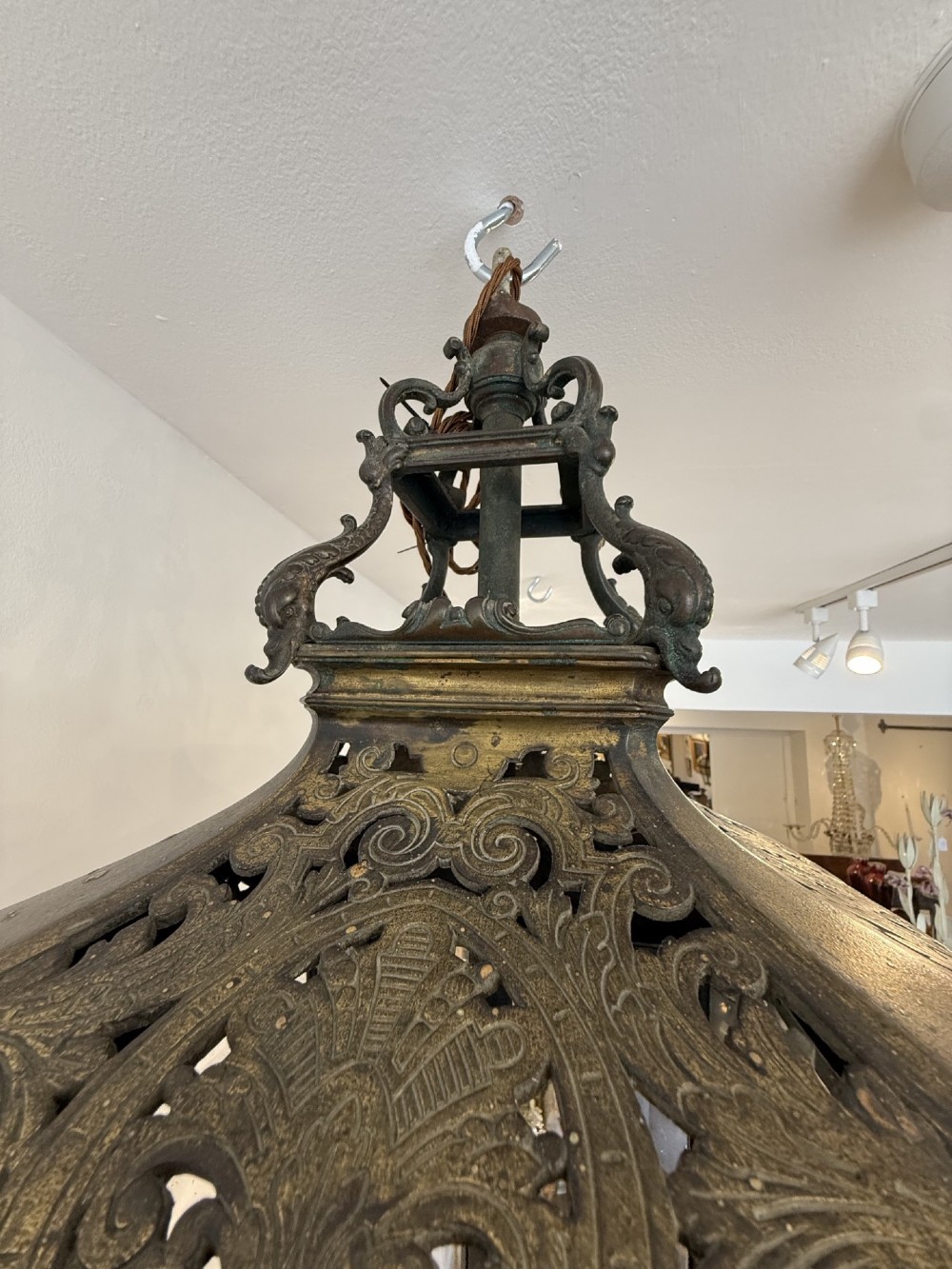 An antique patinated bronze hanging hall lantern featuring clear glass panels, ornate pierced metal cresting, and a scrolled lower finial. View VI