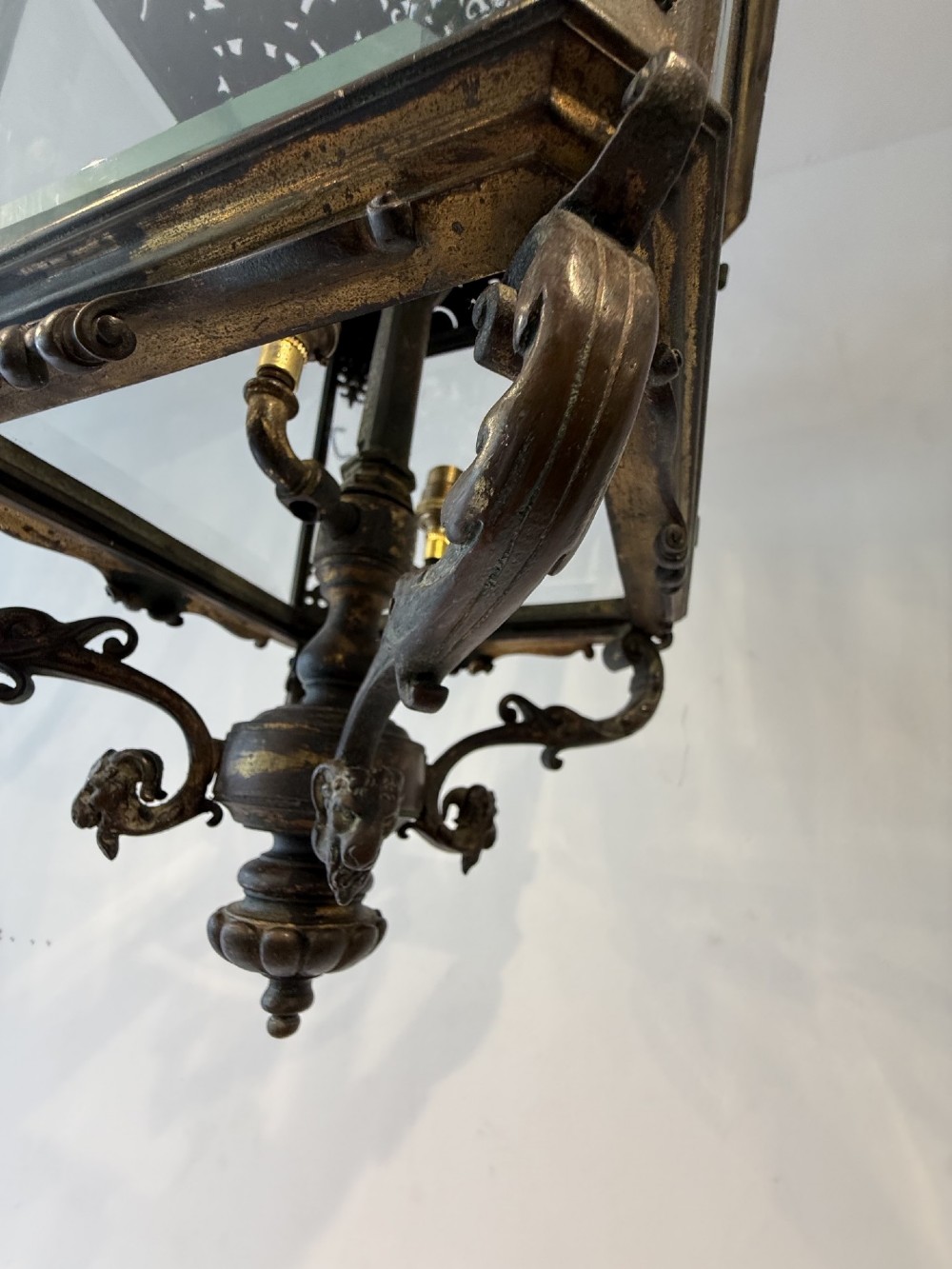 An antique patinated bronze hanging hall lantern featuring clear glass panels, ornate pierced metal cresting, and a scrolled lower finial. View VII