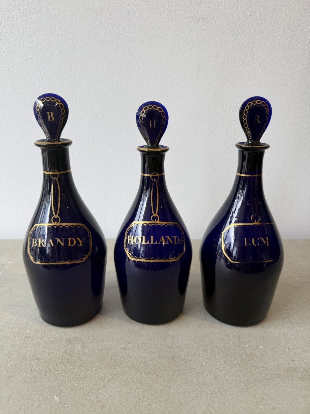 A set of three cobalt blue glass decanters with gilded labels for Brandy, Hollands, and Rum, complete with matching initialled stoppers.