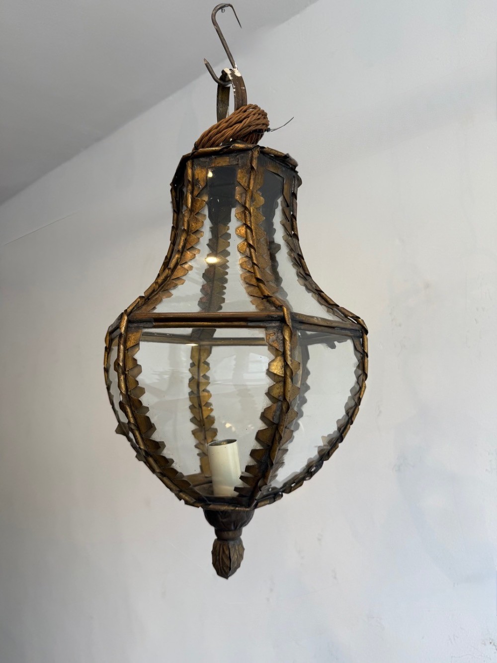 A vintage brass pendant lantern featuring curved glass panels framed with notched brass edging and a decorative bottom finial.