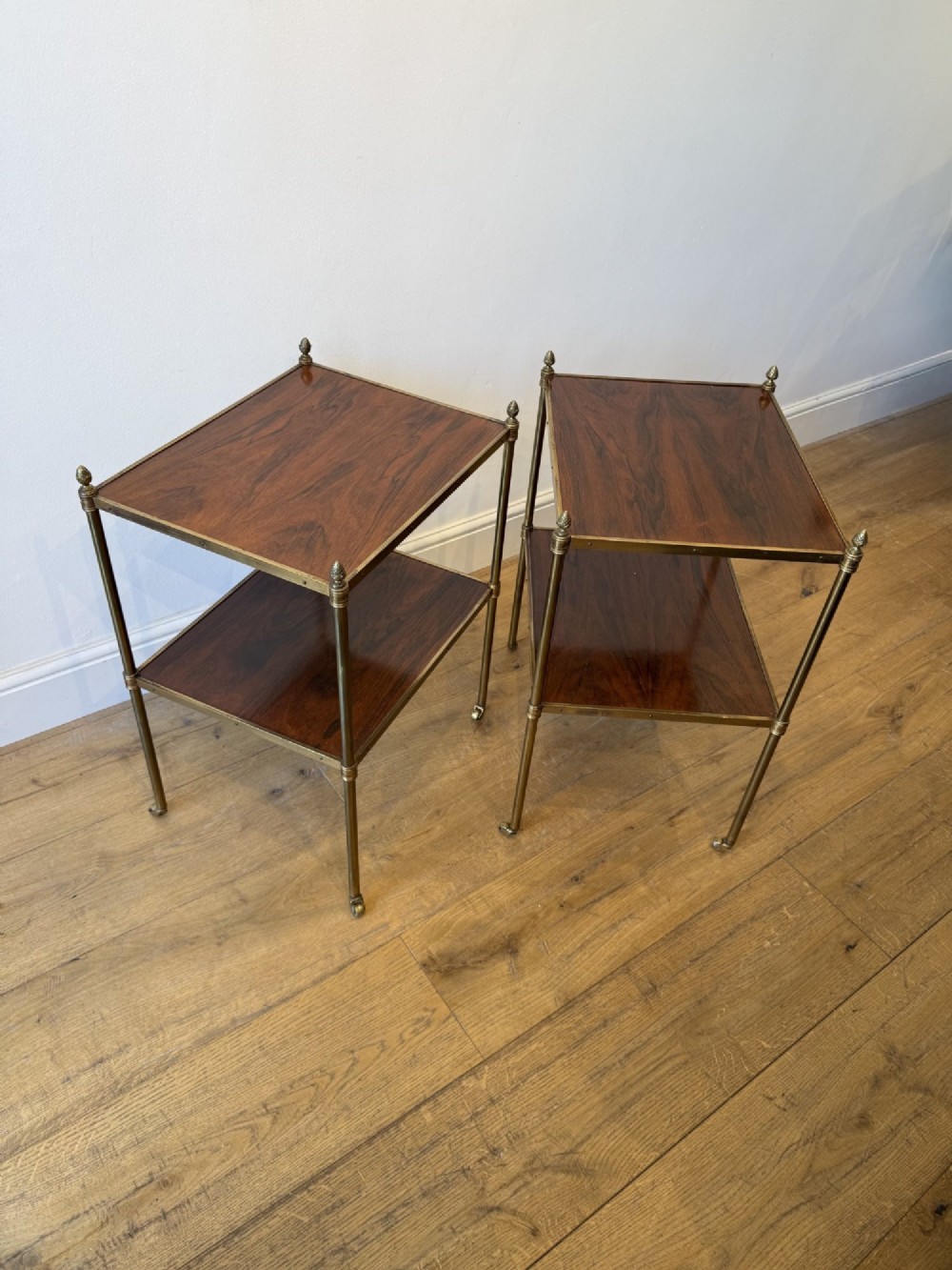 A pair of two-tier brass side tables featuring dark polished wood shelves, finial tops, and slender legs with small castors.