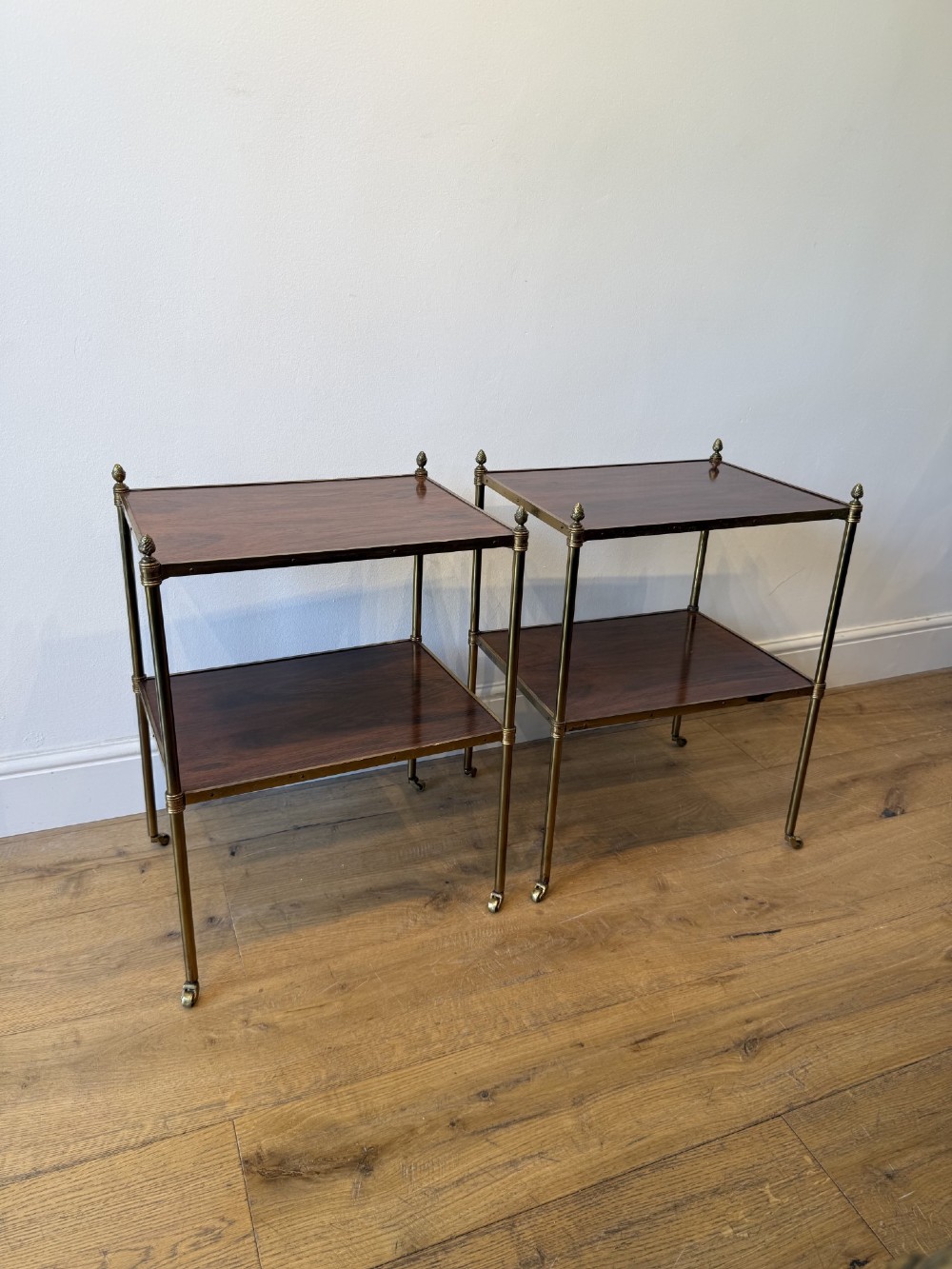 A pair of two-tier brass side tables featuring dark polished wood shelves, finial tops, and slender legs with small castors. View IV