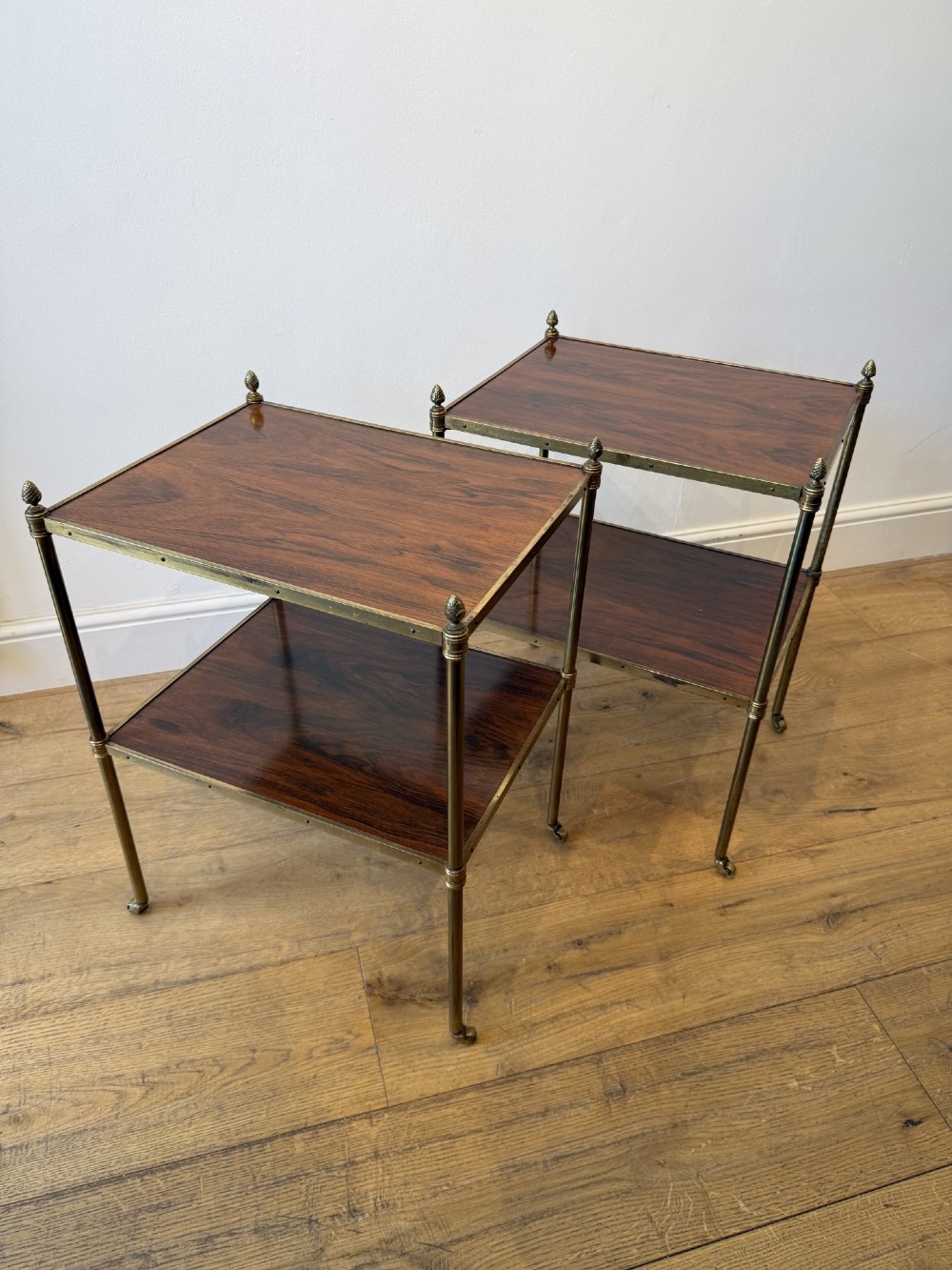 A pair of two-tier brass side tables featuring dark polished wood shelves, finial tops, and slender legs with small castors. View V