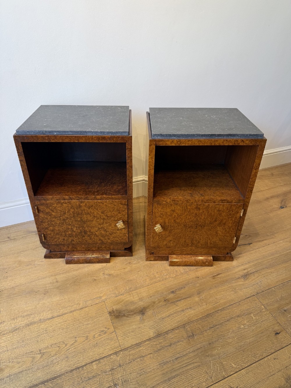 A pair of Art Deco burr walnut bedside cabinets featuring dark stone tops, open upper shelves, and single cupboard doors with square metal handles.