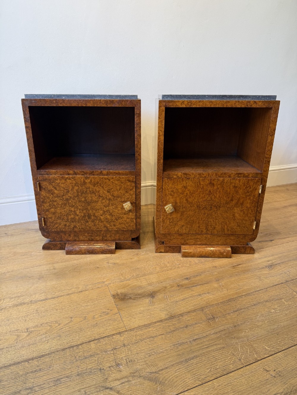 A pair of Art Deco burr walnut bedside cabinets featuring dark stone tops, open upper shelves, and single cupboard doors with square metal handles. View III