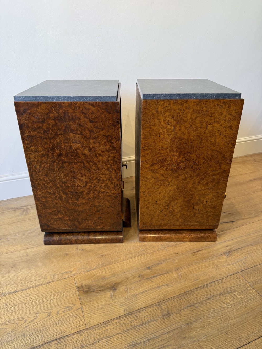 A pair of Art Deco burr walnut bedside cabinets featuring dark stone tops, open upper shelves, and single cupboard doors with square metal handles. View V