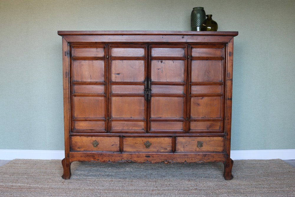 A warm brown wooden Chinese storage cabinet featuring panelled double doors, central brass hardware, three lower drawers, and a carved apron.