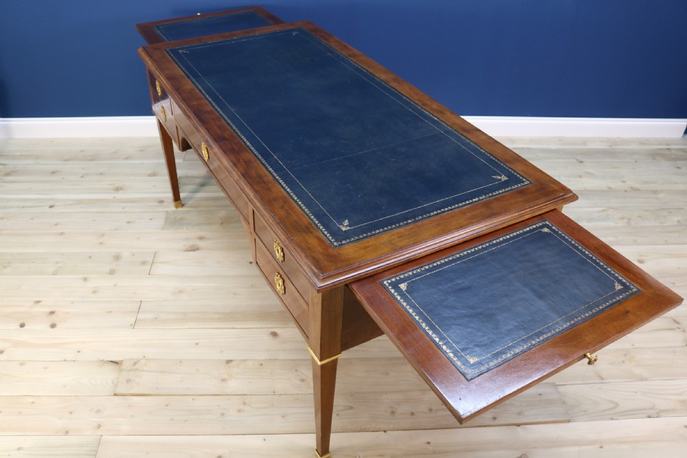 A nineteenth-century Directoire-style wooden bureau plat desk featuring five drawers with brass keyhole escutcheons, tapered legs with brass feet, and a dark leather inset writing top. View XVI