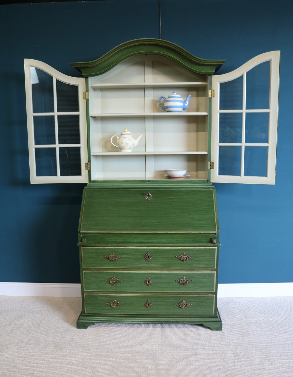 A green painted secretaire writing cabinet featuring an arched glazed upper display section, a fall-front desk revealing fitted compartments, three lower drawers with brass handles, and a matching upholstered chair. View XI