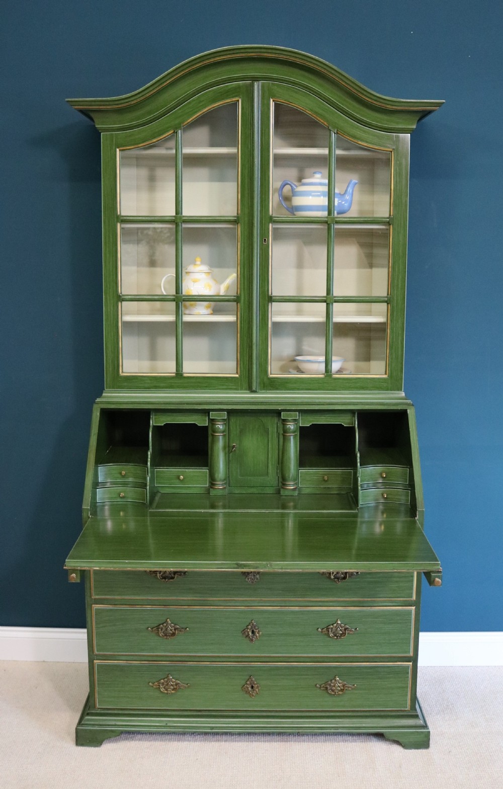 A green painted secretaire writing cabinet featuring an arched glazed upper display section, a fall-front desk revealing fitted compartments, three lower drawers with brass handles, and a matching upholstered chair. View X