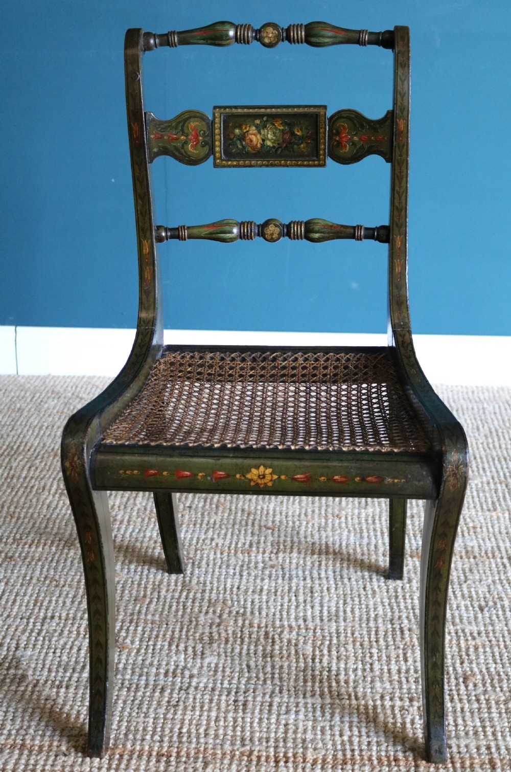 A set of six dark green hand-painted dining chairs featuring sabre legs, turned top rails, painted splats, and woven cane seats. View XII