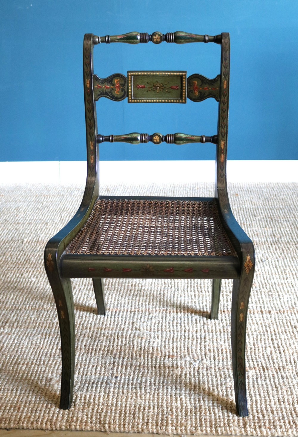 A set of six dark green hand-painted dining chairs featuring sabre legs, turned top rails, painted splats, and woven cane seats. View VI