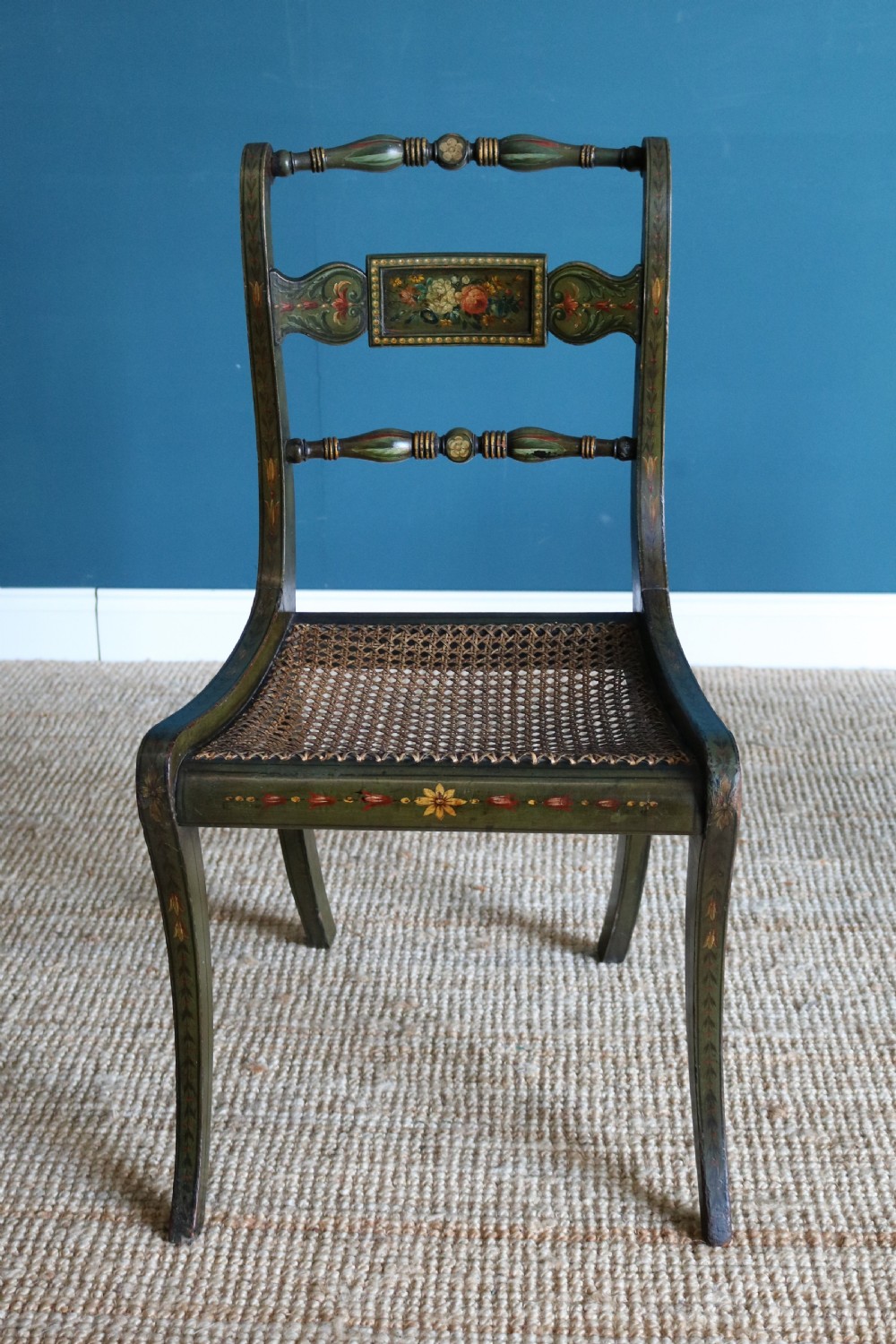 A set of six dark green hand-painted dining chairs featuring sabre legs, turned top rails, painted splats, and woven cane seats. View VII