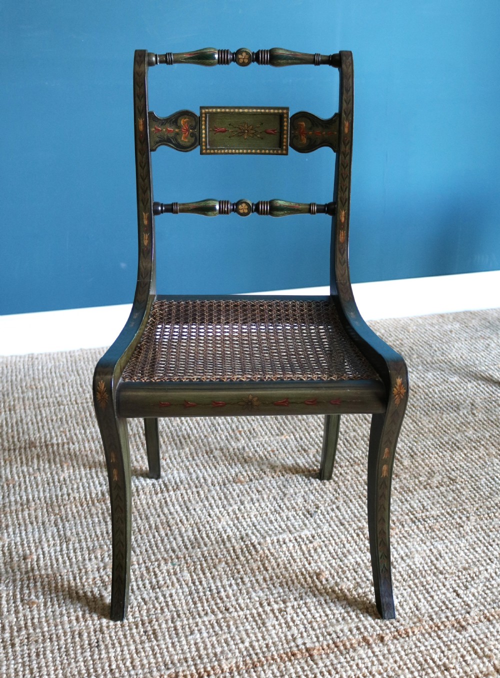 A set of six dark green hand-painted dining chairs featuring sabre legs, turned top rails, painted splats, and woven cane seats. View VIII