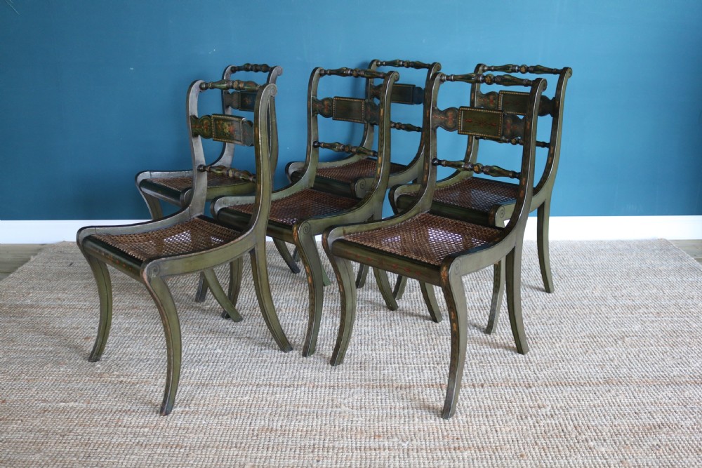 A set of six dark green hand-painted dining chairs featuring sabre legs, turned top rails, painted splats, and woven cane seats. View X