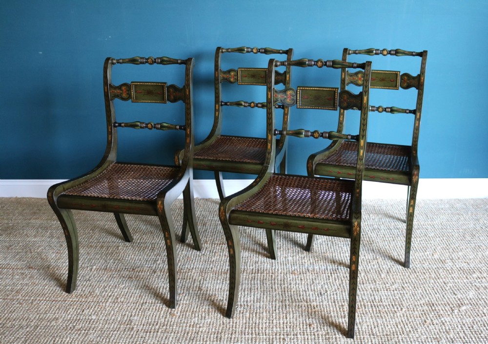 A set of six dark green hand-painted dining chairs featuring sabre legs, turned top rails, painted splats, and woven cane seats. View V