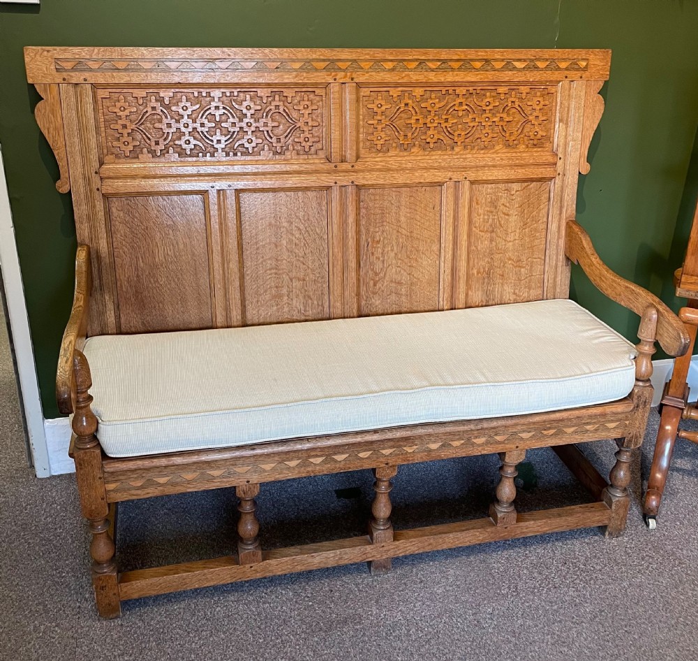 An antique Arts and Crafts solid oak settle featuring a carved high-panel back, turned legs, and a cream upholstered seat cushion.