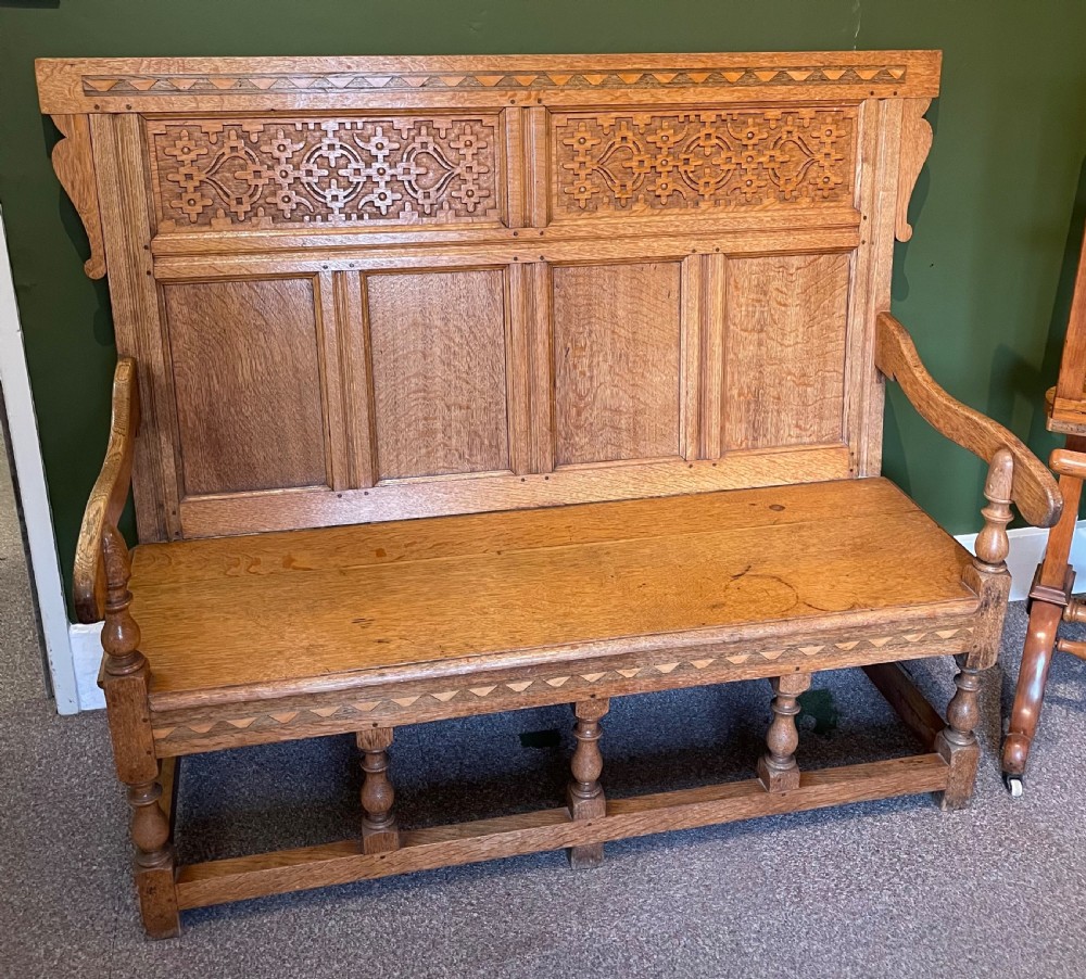 An antique Arts and Crafts solid oak settle featuring a carved high-panel back, turned legs, and a cream upholstered seat cushion. View II