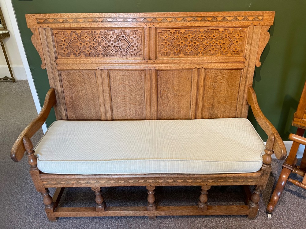 An antique Arts and Crafts solid oak settle featuring a carved high-panel back, turned legs, and a cream upholstered seat cushion. View IV