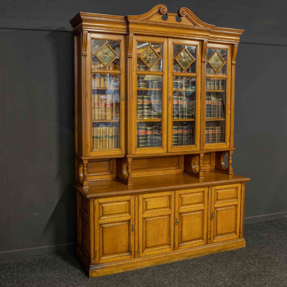 An Edwardian golden oak glazed bookcase featuring a swan-neck pediment, leaded glass doors with stained glass roundels, and a four-door cupboard base.