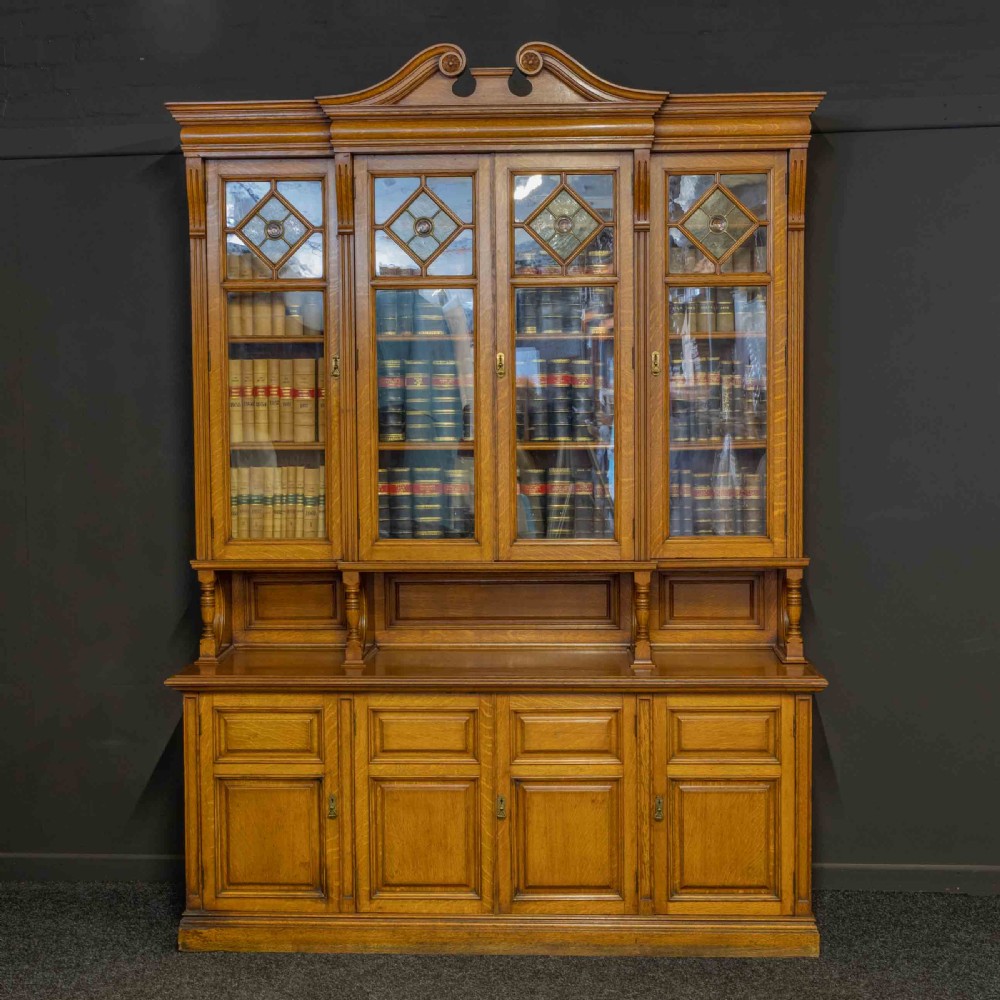 An Edwardian golden oak glazed bookcase featuring a swan-neck pediment, leaded glass doors with stained glass roundels, and a four-door cupboard base. View II