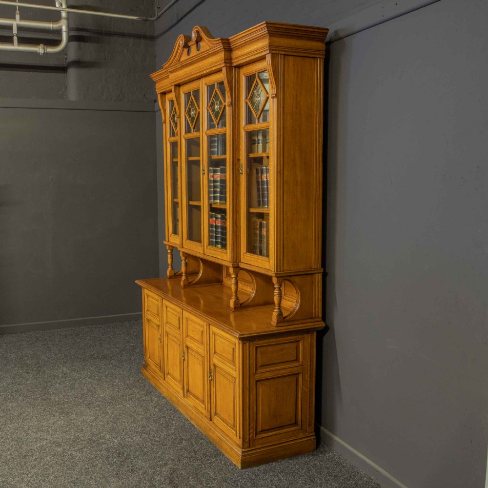 An Edwardian golden oak glazed bookcase featuring a swan-neck pediment, leaded glass doors with stained glass roundels, and a four-door cupboard base. View VI