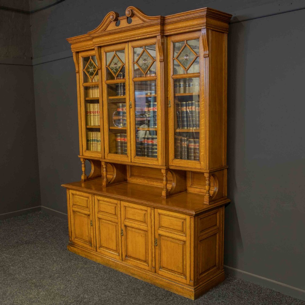 An Edwardian golden oak glazed bookcase featuring a swan-neck pediment, leaded glass doors with stained glass roundels, and a four-door cupboard base. View VII