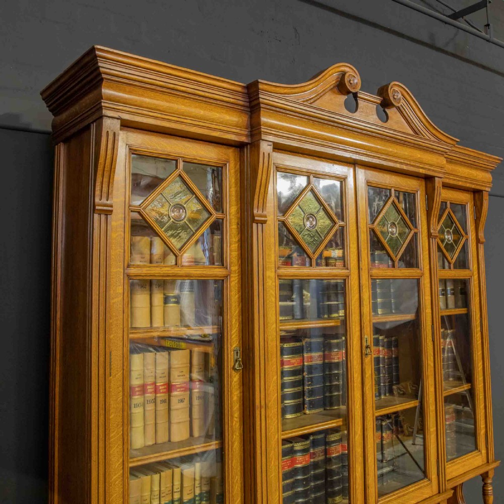 An Edwardian golden oak glazed bookcase featuring a swan-neck pediment, leaded glass doors with stained glass roundels, and a four-door cupboard base. View IX