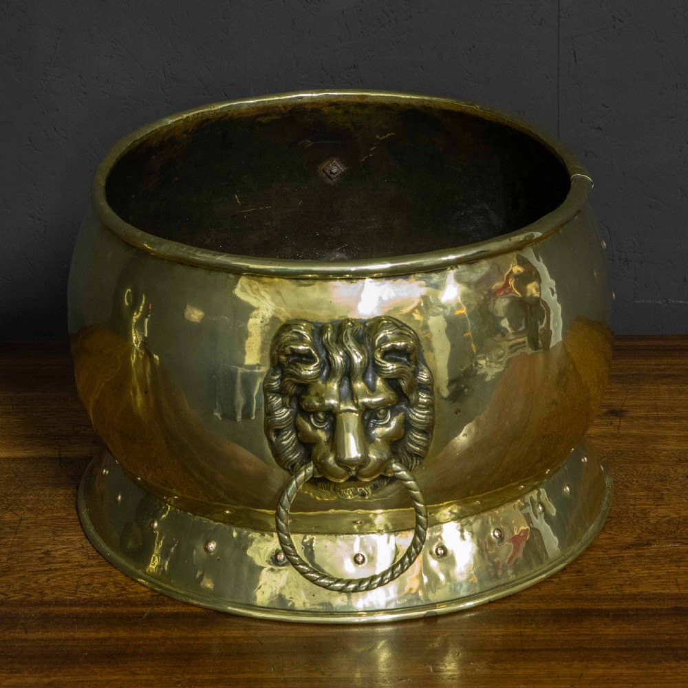 A rounded polished brass vessel with a flared riveted base and cast lion head handles holding rope-twist rings.