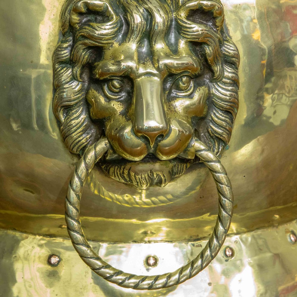A rounded polished brass vessel with a flared riveted base and cast lion head handles holding rope-twist rings. View II