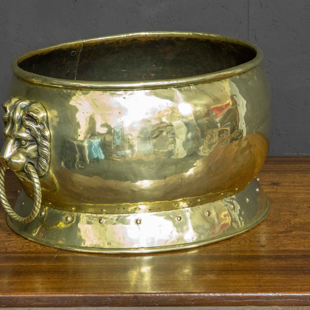 A rounded polished brass vessel with a flared riveted base and cast lion head handles holding rope-twist rings. View III