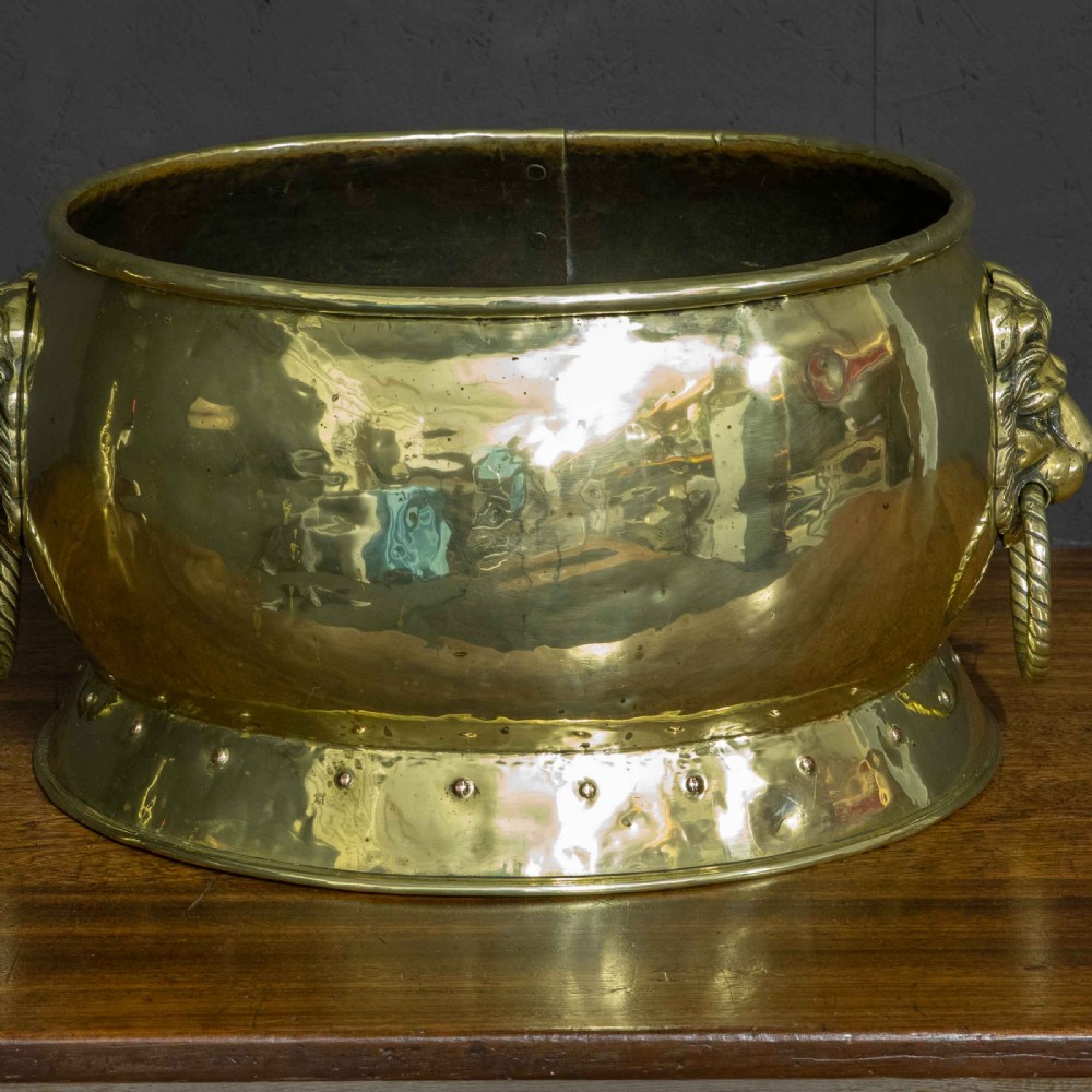 A rounded polished brass vessel with a flared riveted base and cast lion head handles holding rope-twist rings. View IV