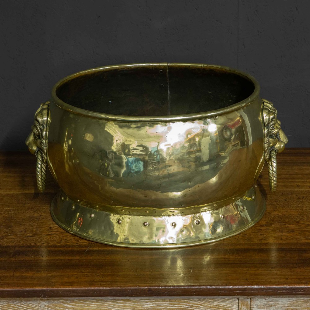 A rounded polished brass vessel with a flared riveted base and cast lion head handles holding rope-twist rings. View V