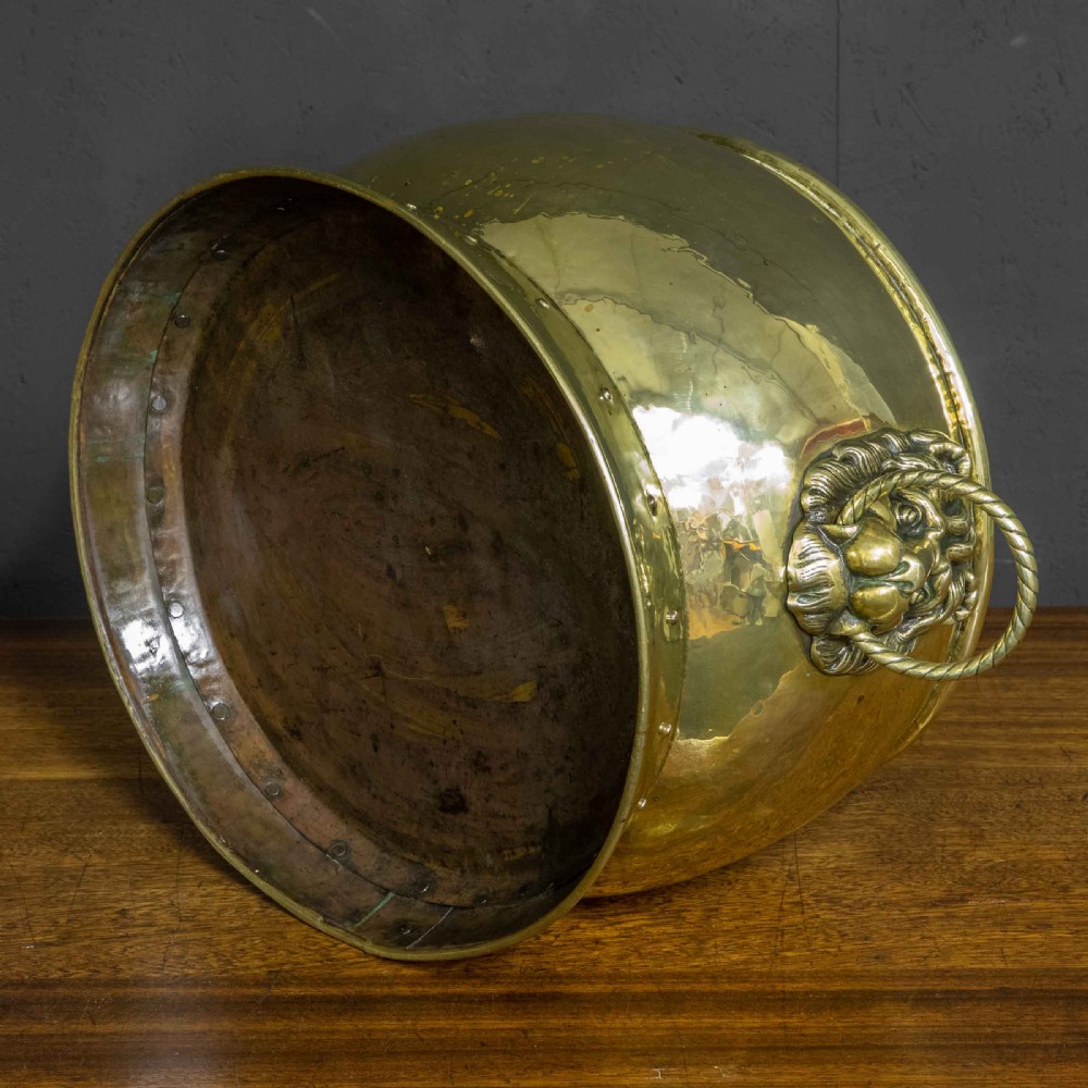 A rounded polished brass vessel with a flared riveted base and cast lion head handles holding rope-twist rings. View VI