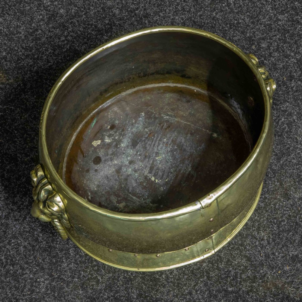A rounded polished brass vessel with a flared riveted base and cast lion head handles holding rope-twist rings. View VII