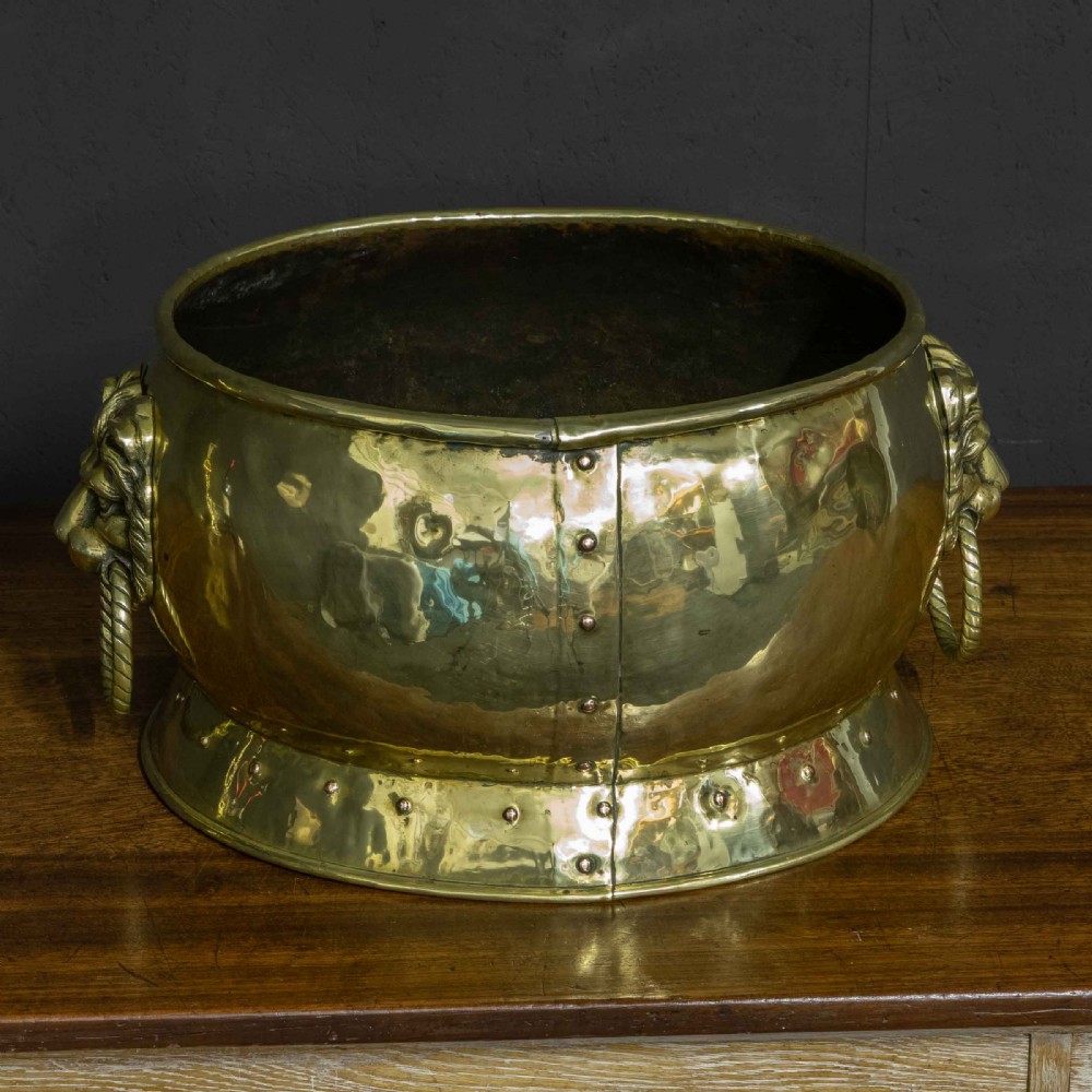 A rounded polished brass vessel with a flared riveted base and cast lion head handles holding rope-twist rings. View VIII