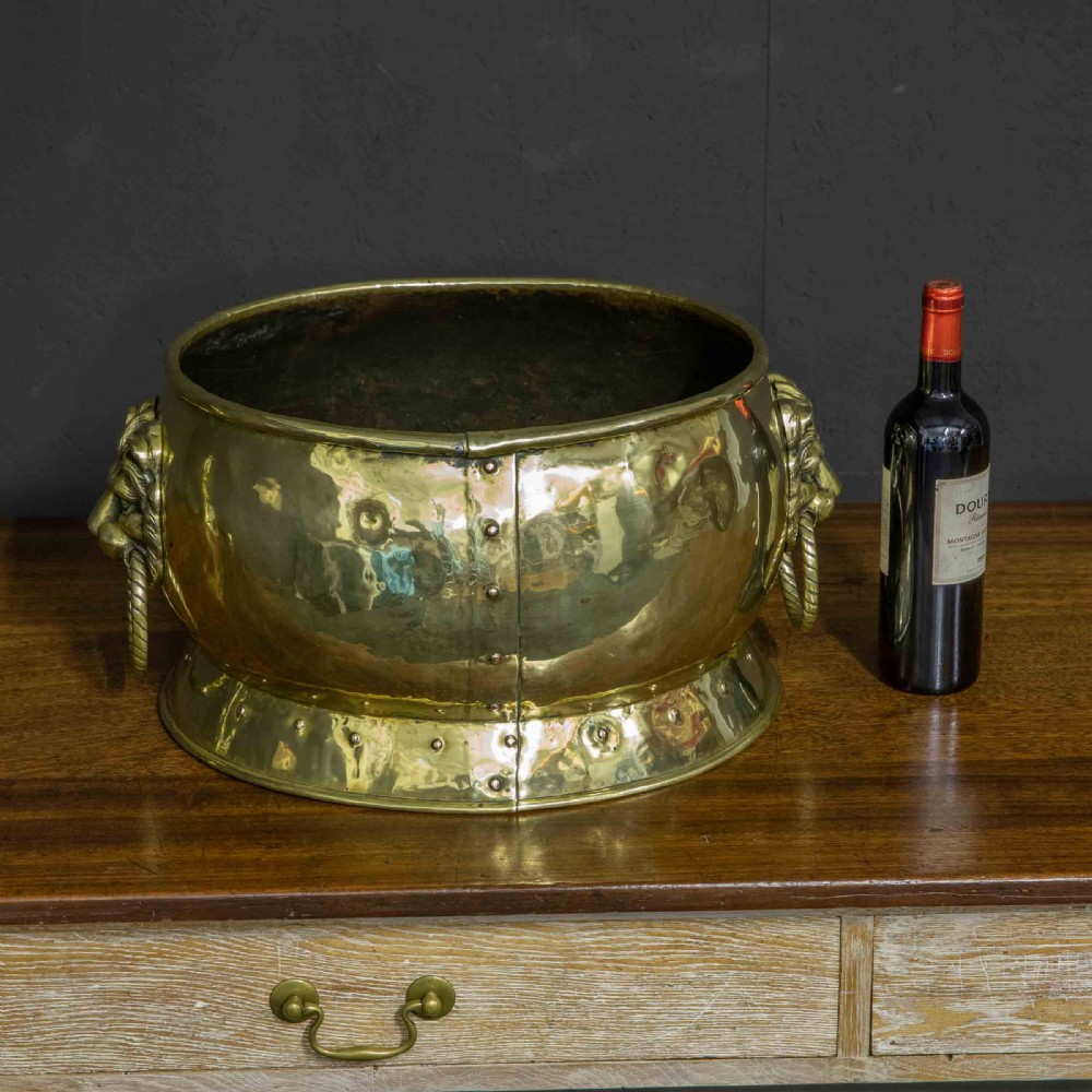A rounded polished brass vessel with a flared riveted base and cast lion head handles holding rope-twist rings. View IX