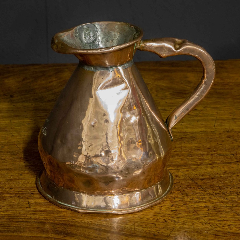 A nineteenth-century copper ale flagon featuring a conical body, a broad curved strap handle, and stamped verification marks inside the lip.