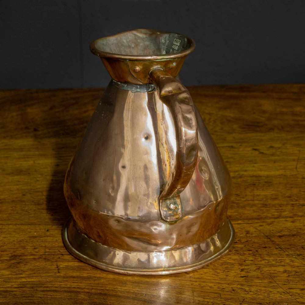 A nineteenth-century copper ale flagon featuring a conical body, a broad curved strap handle, and stamped verification marks inside the lip. View IV