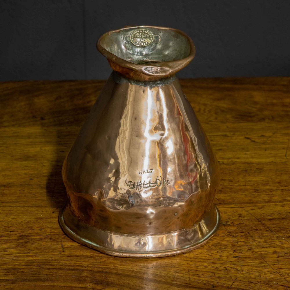 A nineteenth-century copper ale flagon featuring a conical body, a broad curved strap handle, and stamped verification marks inside the lip. View V