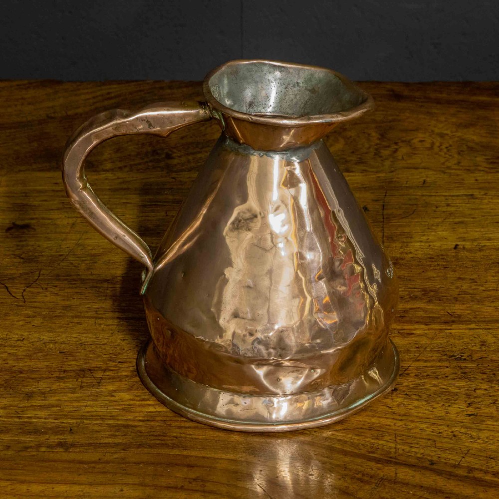 A nineteenth-century copper ale flagon featuring a conical body, a broad curved strap handle, and stamped verification marks inside the lip. View VI