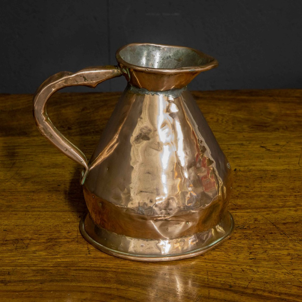 A nineteenth-century copper ale flagon featuring a conical body, a broad curved strap handle, and stamped verification marks inside the lip. View VII